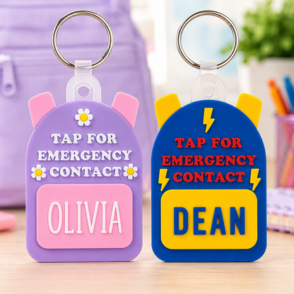 Two colorful emergency contact keychains displayed side by side on a wooden surface. The left keychain is lavender purple with pink ears, white text reading 'TAP FOR EMERGENCY CONTACT' and a pink nameplate reading 'OLIVIA'.