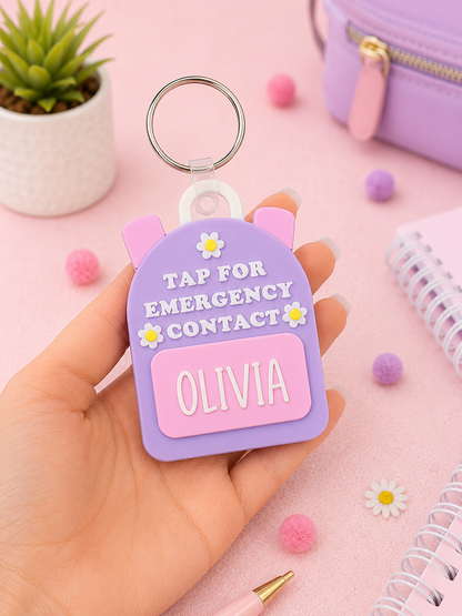 A hand holds a lavender and pink emergency contact keychain shaped like a cat head with the text 'TAP FOR EMERGENCY CONTACT' and a customizable name plate reading 'OLIVIA'. The background is a soft pink desk styled with a small potted succulent, a purple pouch, and stationery.