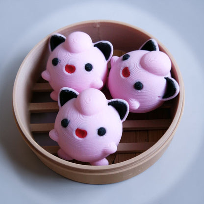 Three jigglypuff toys with black and red details in a wooden steamer basket on a light gray background.